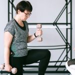 Elderly woman lifting dumbbells with guidance from a trainer indoors.