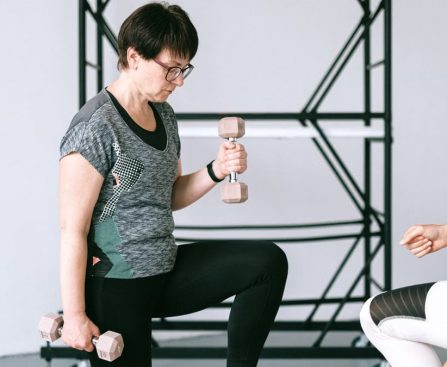 Elderly woman lifting dumbbells with guidance from a trainer indoors.