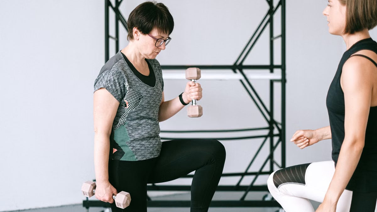 Elderly woman lifting dumbbells with guidance from a trainer indoors.
